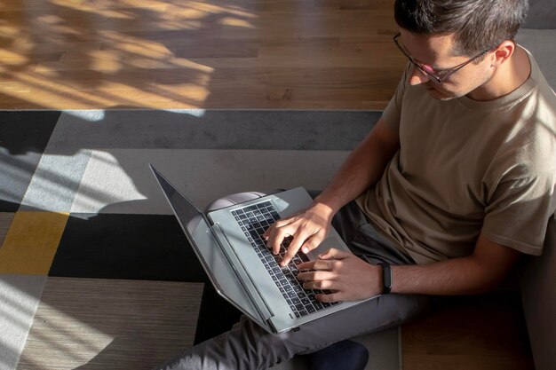 Man in eyeglasses with a laptop is working from home by sitting on the floor in a modern and stylish interior