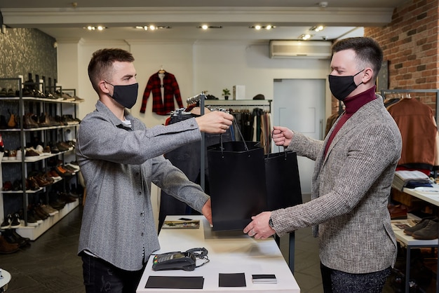 A man in a face mask to avoid the spread of coronavirus is taking his purchase from a seller in a clothing store. A male shop assistant is giving a second bag with clothes to a customer in a boutique