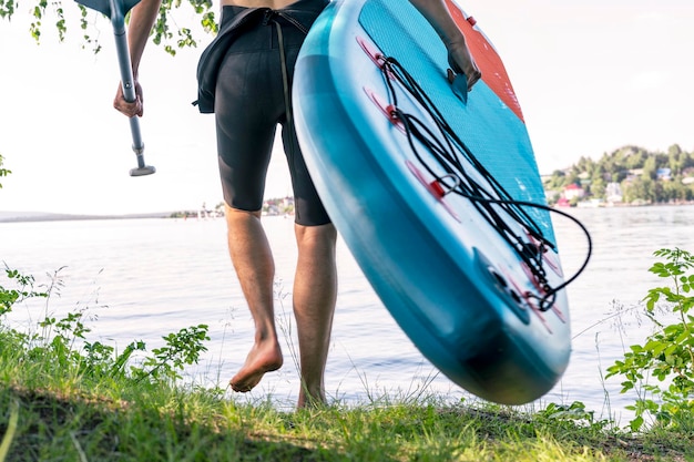 Man faceless in thermo suit with stand up paddleboard blue SUP by shore of lake with paddle male