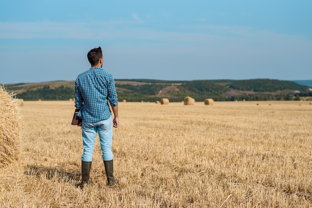 Man farmer agronomist in jeans and shirt stands back in the field after haymaking, with tablet looking into the distance.