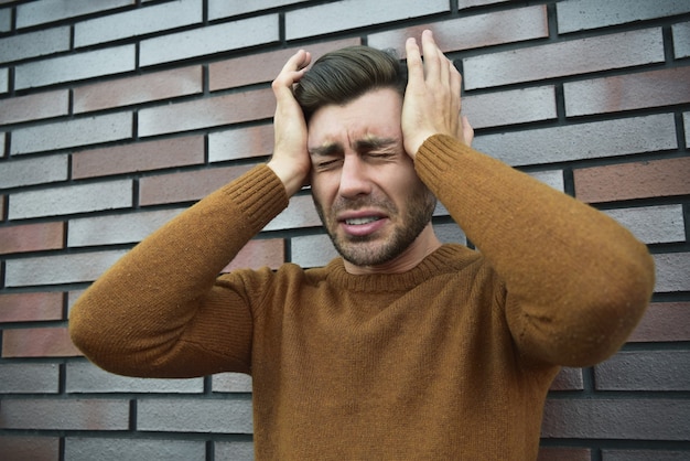 Man feeling painful emotions, screaming, witnessing death of close friend, being miserable and devastated, holding palms on head, yelling from pain and stress, standing unhappy over brown brick wall