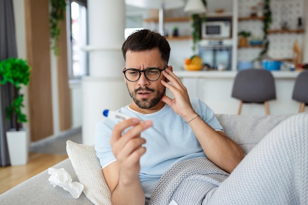 Man feeling sick lying in the bed and looking the thermometer\
sick man lying on sofa checking his temperature under a blanket at\
home in the living room