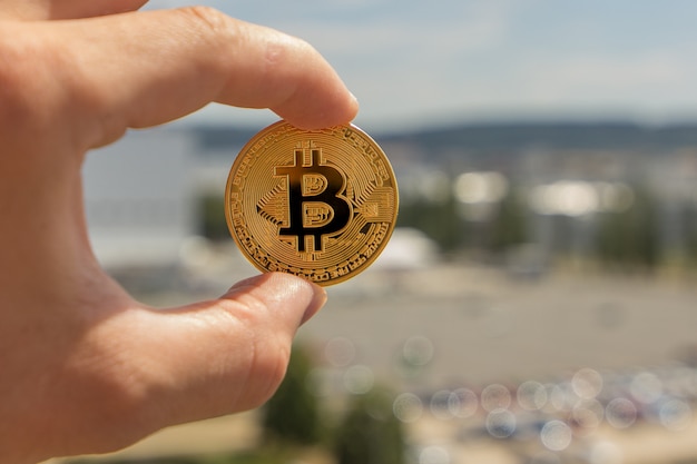 Man fingers are holding a big round golden bitcoin coin in front of industrial city.