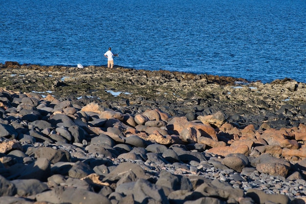 Man fishing in the atlantic ocean on the rough terrain beach\
formed by cooled lava volcanic rocks