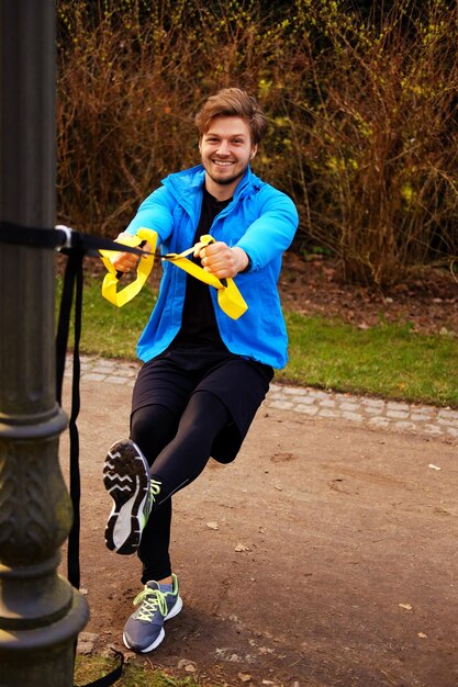 Man fitness-model in een blauwe sportjas die trainingen doet met fitness trx-strips in een park.