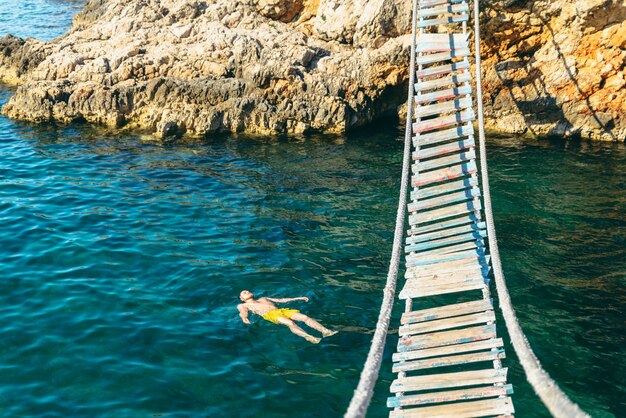 Man floating in sea water on back. bay with rocky beach and suspension bridge. copy space. summer vacation