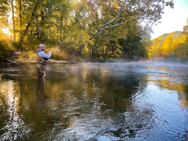 Мужчина ловит рыбу нахлыстом в реке с осенними цветами на заднем плане.