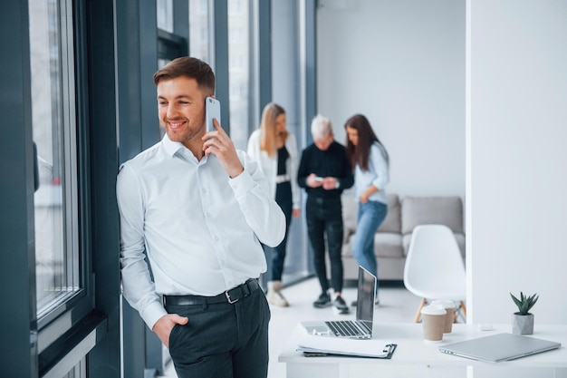 Man in formal clothes talking by phone and standing in front of young successful team that working and communicating together indoors in office