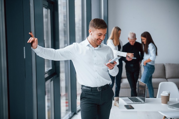 Man in formal clothes with phone and in headphones standing in front of young successful team that working and communicating together indoors in office