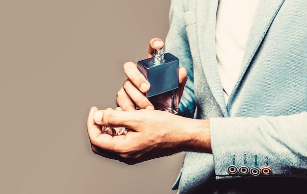 Man in formal suit, bottle of perfume, closeup. Fragrance smell. Men perfumes. Fashion cologne bottle. Man holding up bottle of perfume. Men perfume in the hand on suit background. Copy space.