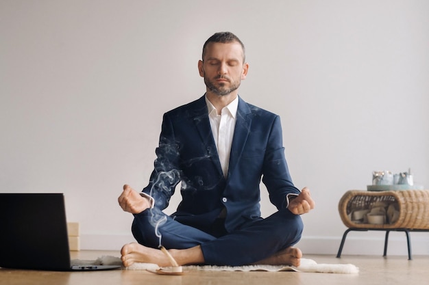 A man in a formal suit meditates while sitting in a fitness room with a laptop