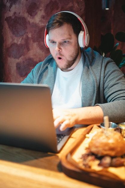 Man freelancer in headset working on laptop in cafe eating burger and drinking tea copy space