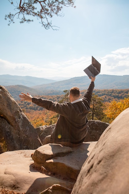 Man freelancer werkt op laptop op de top van de rots met prachtig uitzicht op herfstbos