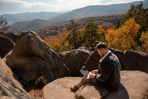 Мужчина-фрилансер работает на ноутбуке на вершине скалы с прекрасным видом на осенний лес