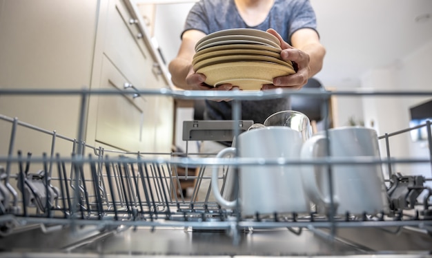 A man in front of an open dishwasher takes out or puts down dishes.