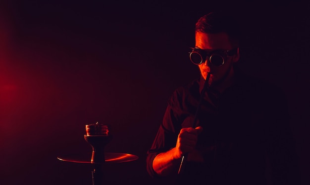 Man in futuristic glasses smokes a hookah in a bar with red neon lights