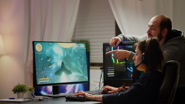 Uomo giocatore che insegna alla sua ragazza a giocare a un videogioco sparatutto spaziale su un potente personal computer rgb. pro cyber donna con auricolare che esegue videogiochi in streaming da casa durante il torneo online