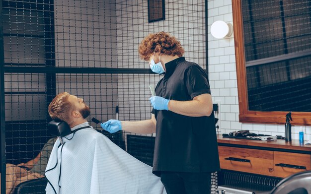 Man getting hair cut at the barbershop wearing mask during coronavirus pandemic. Professional barber wearing gloves. Covid-19, beauty, selfcare, style, healthcare and medicine concept.