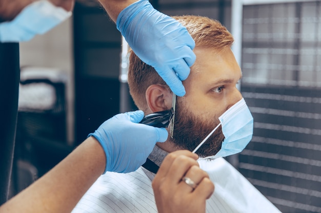 Man getting hair cut at the barbershop wearing mask during coronavirus pandemic. Professional barber wearing gloves. Covid-19, beauty, selfcare, style, healthcare and medicine concept.