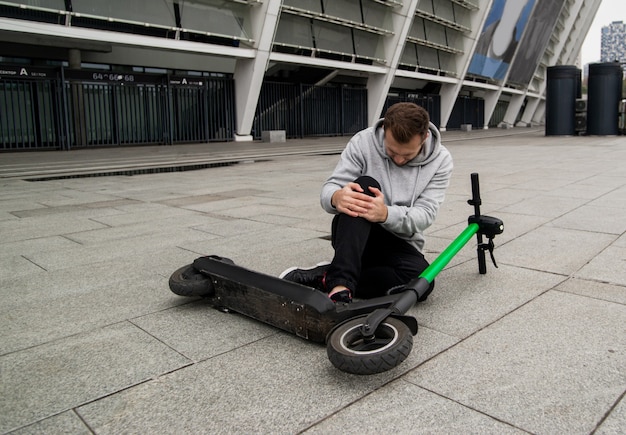 Man gevallen van scooter terwijl hij zijn knie vasthield en pijn voelde. Groene elektrische scooter liggend op asfalt. Stijlvolle man in grijze hoody zit op de grond en heeft kniepijn. Milieuvriendelijk transportconcept.