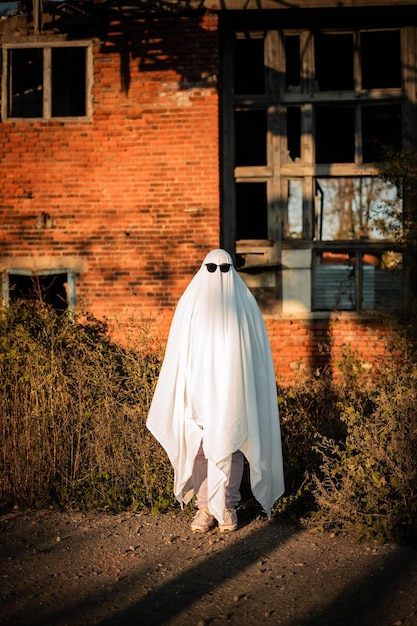 Foto un uomo in un costume di fantasma fatto di un lenzuolo e occhiali da sole si trova vicino a un edificio abbandonato ghost challenge 2021 sta festeggiando halloween