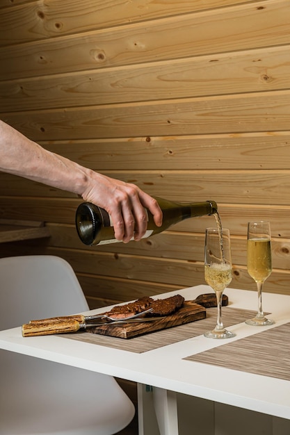 Man gieten witte wijn uit een fles in een fluitglas gekookt en gesneden steak vlees geserveerd op houten snijplank met bestek op tafel