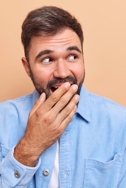 man giggles and cons mouth with hand tries to hide his emotions laughs at something funny wears blue velvet shirt isolated on beige