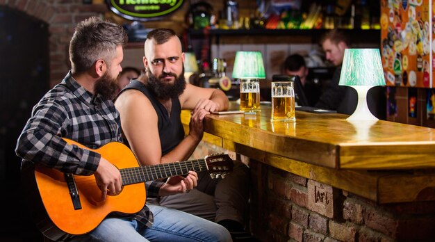 Man gitaar spelen in bar Vrolijke vrienden ontspannen met gitaarmuziek Vrijdag ontspanning in bar Vrienden ontspannen in bar of pub Echte mannen vrije tijd Hipster brutale bebaarde vrije tijd doorbrengen met vriend in bar