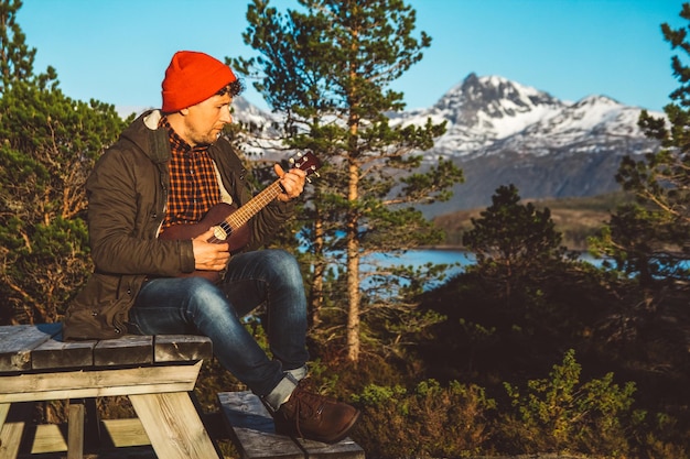 Man gitaarspelen zittend op een houten tafel tegen de achtergrond van bergen, bossen en meren