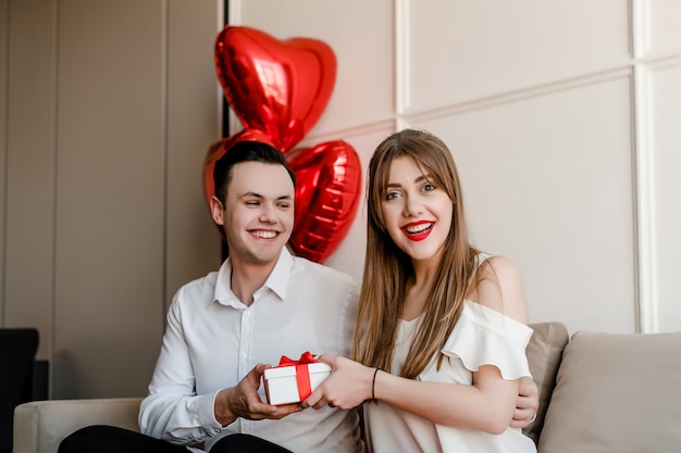 Мужчина дарит подарок улыбающейся женщине с воздушными шарами в форме сердца на диване у себя дома