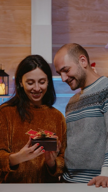 お祝いの装飾が施されたキッチンでクリスマスのお祝いのために女性にプレゼントを与える男性。冬の休日のお祭りのパートナーからの贈り物を受け取って驚いた人。陽気なカップル