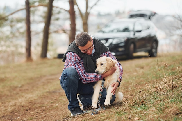 眼鏡をかけた男は、森の中で彼の犬と一緒に屋外で散歩をしています現代の黒い車の後ろ