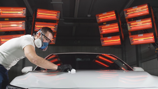 Man in glasses and a mask, a respirator polishes the car with a sponge and a special chemical composition to protect the paint on the body from scratches