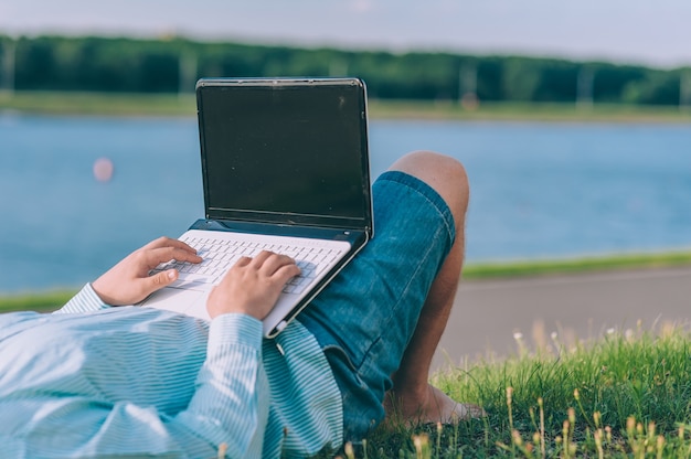 A man in glasses and a shirt working on a laptop in the fresh air, against the space of a river.