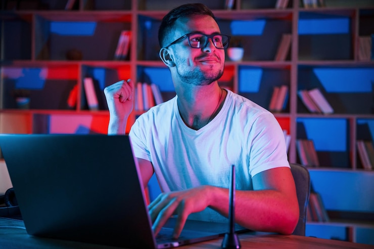 Photo man in glasses and white shirt is sitting by the laptop in dark room with neon lighting