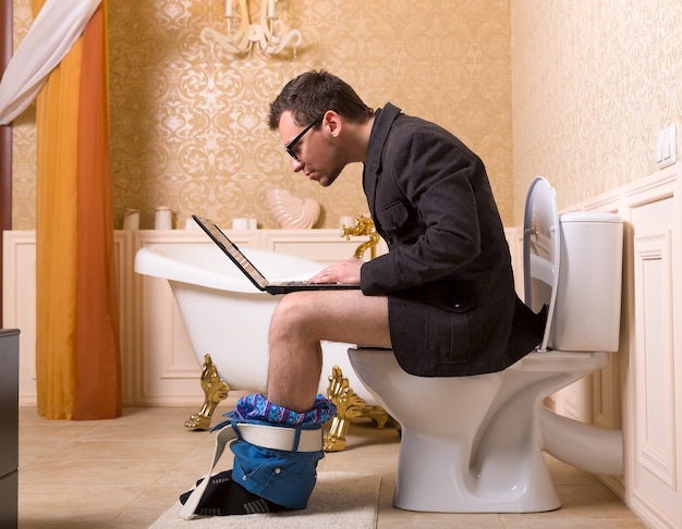 Man in glasses with laptop sitting on the toilet bowl. Luxury bathroom interior in vintage style 