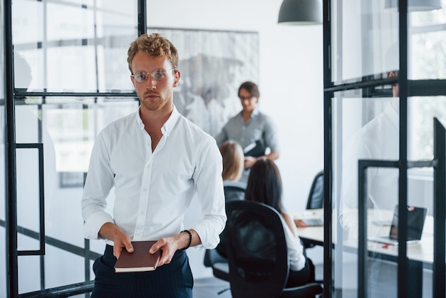 Man in glasses with notepad in hands stands in the office opposite employees.