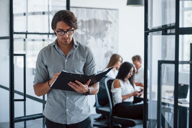 Man in glasses with notepad and pen in hands stands in the office opposite employees.