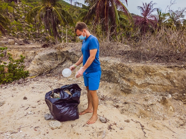 장갑을 낀 남자는 바다를 오염시키는 비닐 봉지를 집어들다 인공 오염으로 인한 해변 모래에 엎질러진 쓰레기 쓰레기 문제와 자원 봉사자를 개념적으로 청소하기 위한 환경 캠페인