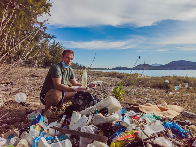 手袋をはめた男が海や森を汚染するプラスチックを拾う人為的な汚染キャンペーンによって引き起こされた砂浜にゴミがこぼれるという問題