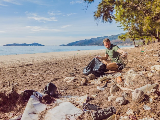 手袋をはめた男が海や森を汚染するプラスチックを拾う人為的な汚染キャンペーンによって引き起こされた砂浜にゴミがこぼれるという問題