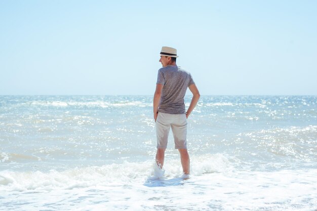 Мужчина в серой рубашке, белых шортах и соломенной шляпе стоит в море