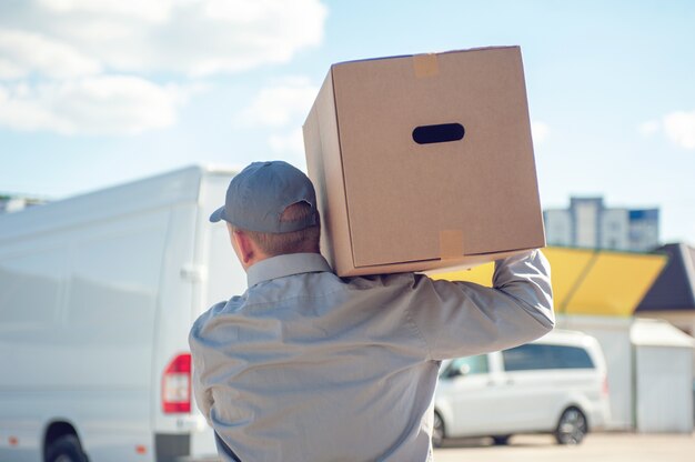 A man in a gray suit in his hands with a cardboard box against the space of the van