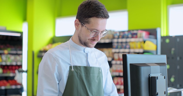 Man in green apron as a cashier at the cash register in the supermarket or discounter Handsome man smiling to the camera