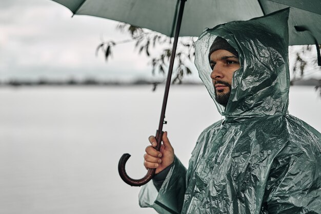 Un uomo in un impermeabile verde e con un ombrello sulla riva vicino all'acqua.