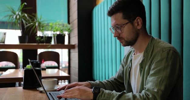 카페나 코워킹 스페이스의 테이블에 앉아 노트북 컴퓨터를 사용하는 녹색 셔츠를 입은 남자 노트북 작업을 하고 카페에서 커피를 마시는 남자