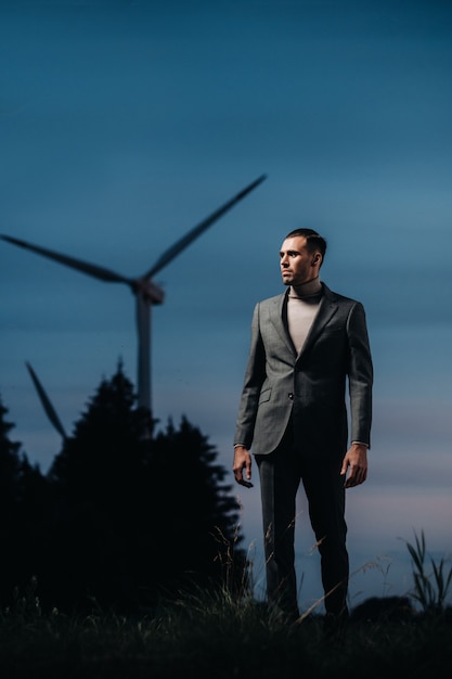 Мужчина в сером деловом костюме стоит рядом с ветряной мельницей после заката. Бизнесмен возле ветряных мельниц в ночное время. Современная концепция будущего.