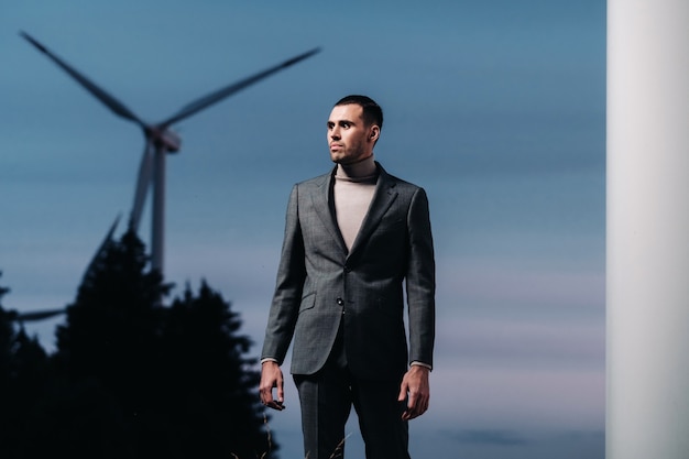 A man in a grey business suit stands next to a windmill after sunset .Businessman near windmills at night.Modern concept of the future.