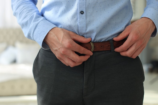 A man in grey pants and a blue shirt buttoned a brown leather trouser belt.