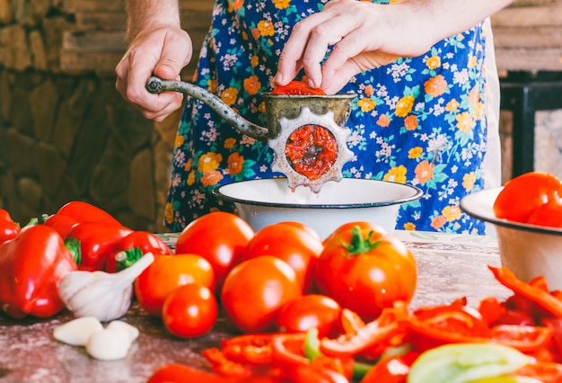 Человек измельчает кусочки спелых помидоров в старинную мясорубку.